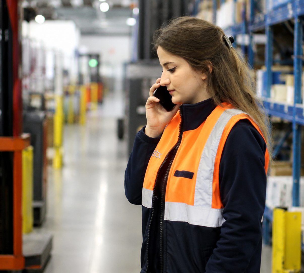 Une salariée en gilet haute visibilité téléphone dans l'allée d'un entrepôt logistique
