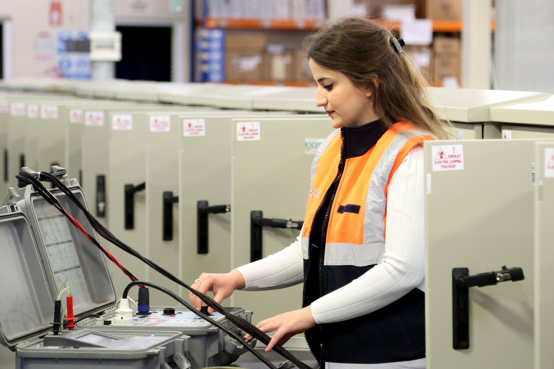 Une technicienne en gilet haute visibilité contrôle des armoires électriques en atelier